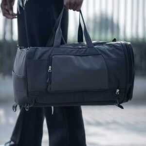 Black duffel bag with multiple compartments being carried by a person in black pants.